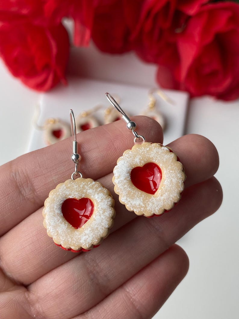May include: A pair of dangle earrings resembling heart-shaped jammy dodgers. The earrings feature a beige biscuit base, a red heart centre, and a dusting of white powder. They hang from silver hooks.