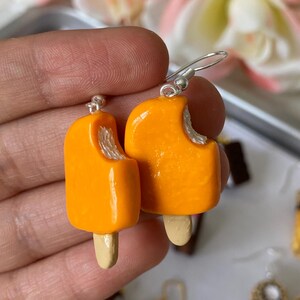 Puede incluir: Un par de pendientes de paletas de naranja con una mordida en cada uno. Los pendientes tienen un gancho plateado y un palito de madera. Un accesorio divertido y juguet&oacute;n.