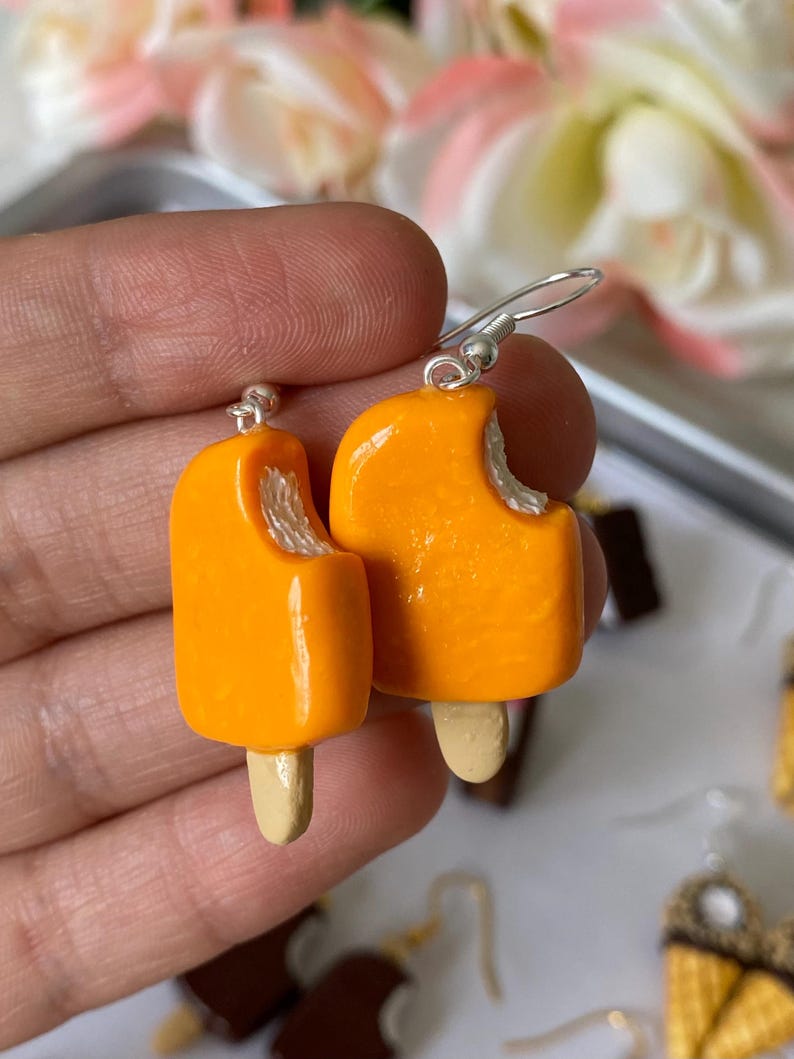 Puede incluir: Un par de pendientes de paletas de naranja con una mordida en cada uno. Los pendientes tienen un gancho plateado y un palito de madera. Un accesorio divertido y juguet&oacute;n.