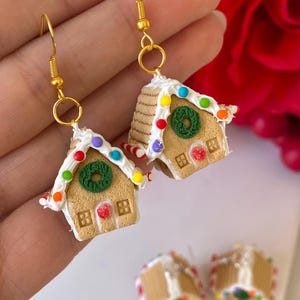 May include: Dangle earrings with a gold-coloured finish, shaped like miniature gingerbread houses. The houses have tan walls, white icing, colourful candy decorations, and a green wreath. Festive earrings, perfect for the holiday season.