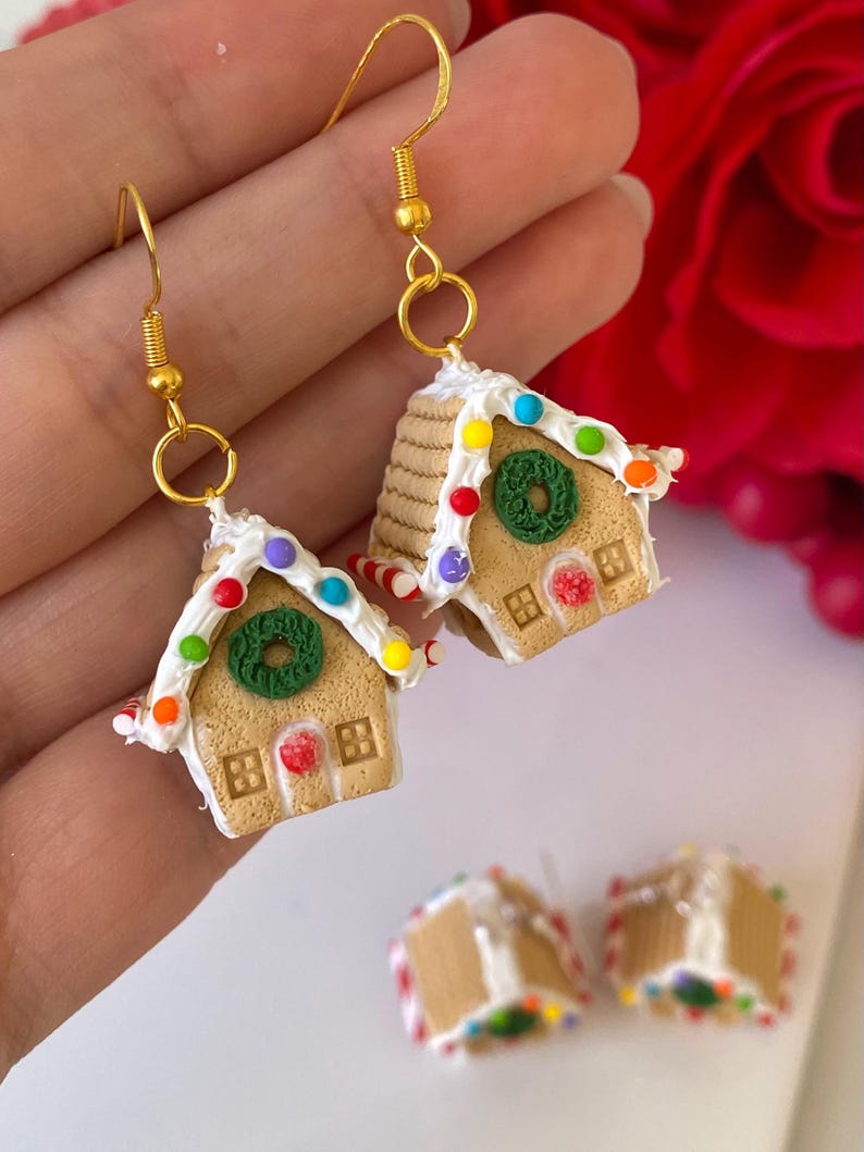 May include: Dangle earrings with a gold-coloured finish, shaped like miniature gingerbread houses. The houses have tan walls, white icing, colourful candy decorations, and a green wreath. Festive earrings, perfect for the holiday season.