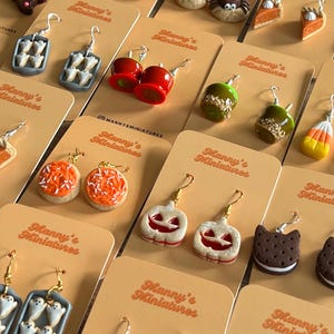 May include: A selection of Halloween-themed earrings, such as spider donuts, pumpkin cookies, and candy apples. The earrings are displayed on tan cards, each bearing the text "Manny's Miniatures".