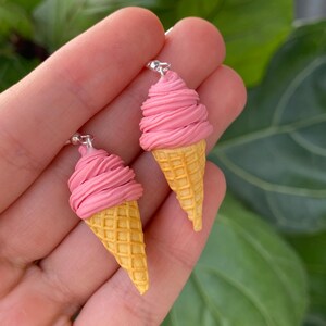 Puede incluir: Un par de pendientes originales con forma de conos de helado. Cada pendiente presenta una parte superior de helado rosa en espiral y una base de cono de gofre amarillo. Los pendientes est&aacute;n suspendidos de ganchos plateados.