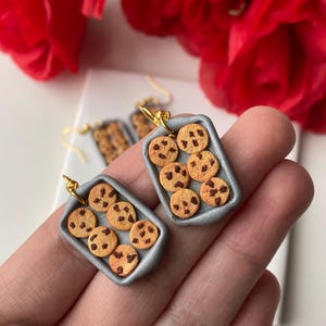 May include: Handmade cookie earrings featuring miniature chocolate chip cookies arranged in a silver baking pan design. The earrings have gold-coloured hooks. The background includes red flowers.