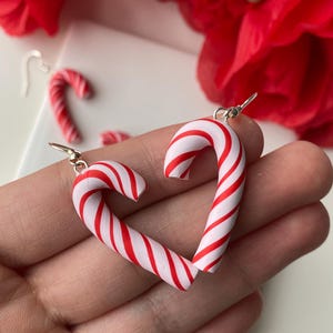 May include: A pair of candy cane earrings in a heart shape, with red and white stripes. The earrings are made from polymer clay and have silver-coloured ear wires. Red flowers are visible in the background.