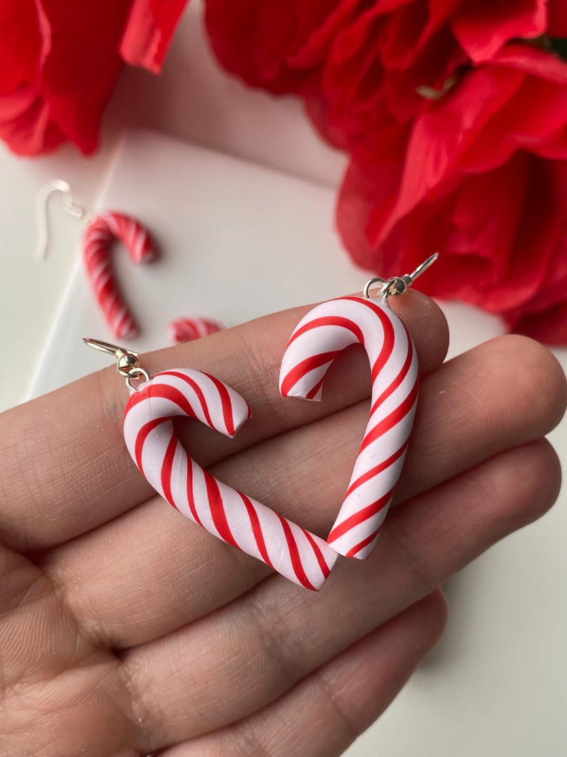 May include: A pair of candy cane earrings in a heart shape, with red and white stripes. The earrings are made from polymer clay and have silver-coloured ear wires. Red flowers are visible in the background.