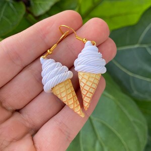 Puede incluir: Un par de pendientes colgantes dorados con forma de conos de helado en miniatura. Cada pendiente tiene un cono amarillo rematado con un remolino de helado blanco. Los pendientes se muestran sobre un fondo verde con hojas.