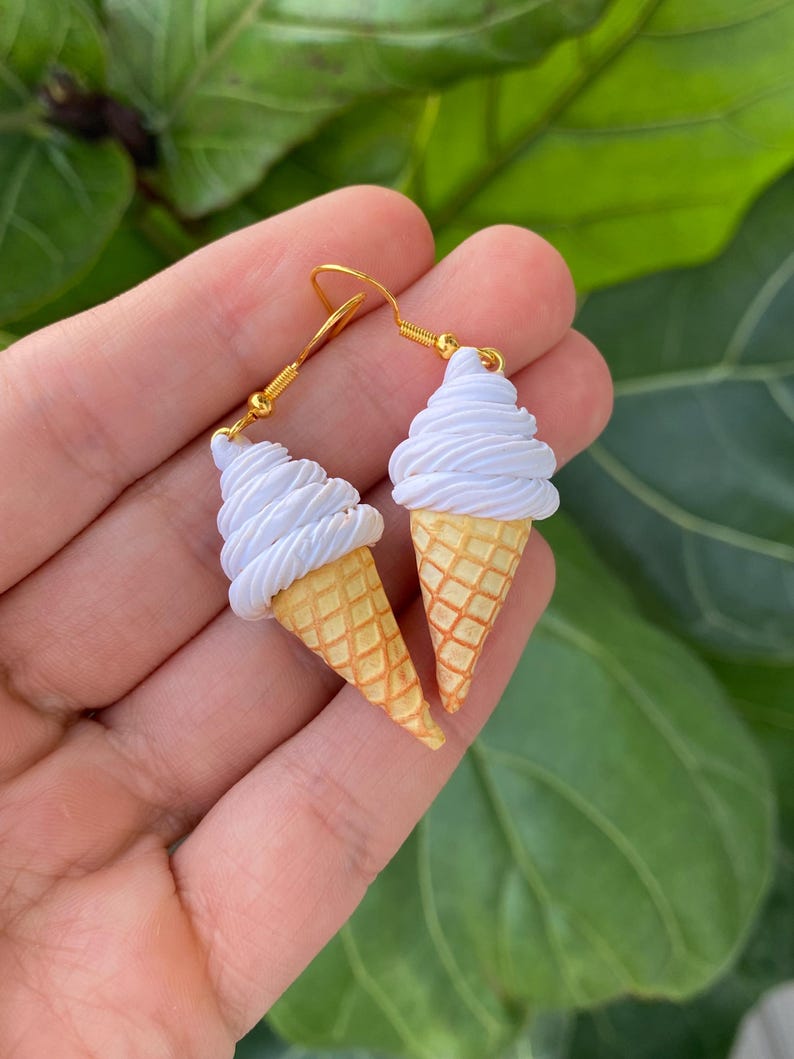 Puede incluir: Un par de pendientes colgantes dorados con forma de conos de helado en miniatura. Cada pendiente tiene un cono amarillo rematado con un remolino de helado blanco. Los pendientes se muestran sobre un fondo verde con hojas.
