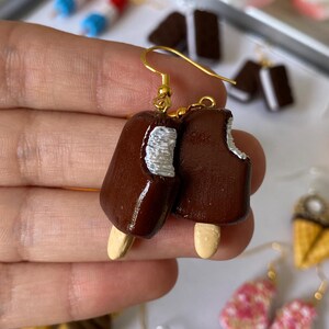 Puede incluir: Pendientes de paletas marrones con ganchos dorados. Uno de los pendientes tiene una mordida. Otros pendientes en el fondo incluyen conos de helado y paletas.