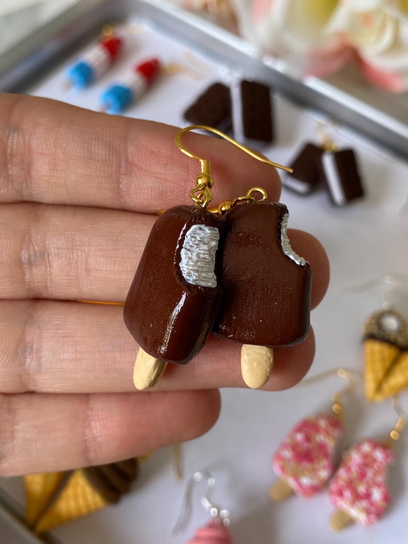 Puede incluir: Pendientes de paletas marrones con ganchos dorados. Uno de los pendientes tiene una mordida. Otros pendientes en el fondo incluyen conos de helado y paletas.