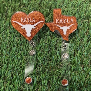 Puede incluir: Dos carretes de insignia naranja brillante, uno en forma de corazón y otro en forma de Texas. Ambos tienen el nombre "Kayla" y un logotipo de cuerno largo blanco. Los carretes tienen correas de plástico transparente y detalles de purpurina naranja.