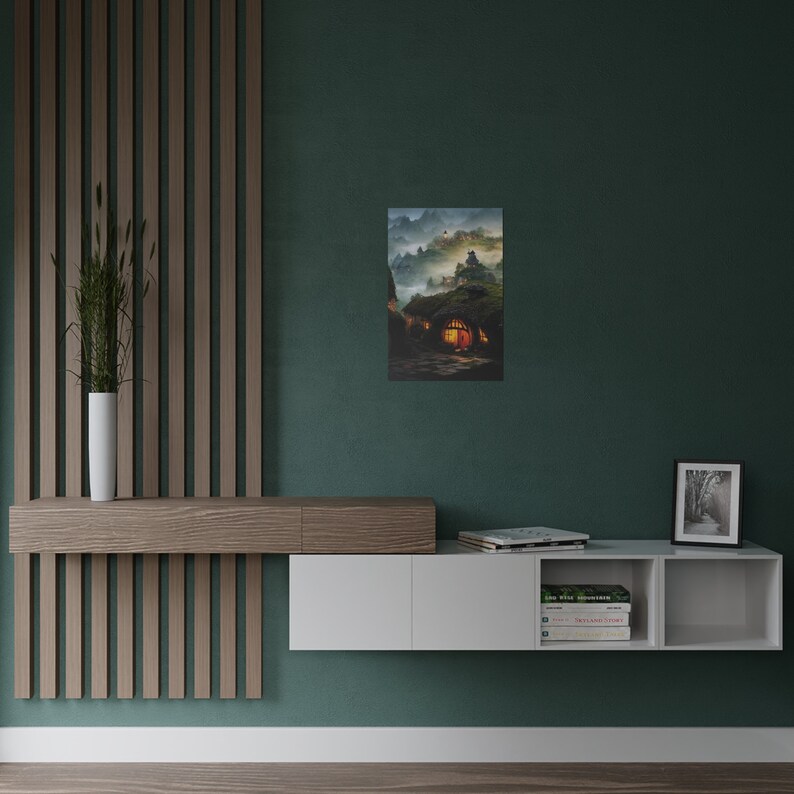 May include: A framed art print depicts a fantasy landscape with a hobbit-hole style dwelling. The artwork features a warm, inviting glow emanating from the windows of the home, set against a backdrop of misty mountains and a twilight sky. The print is displayed on a teal wall.