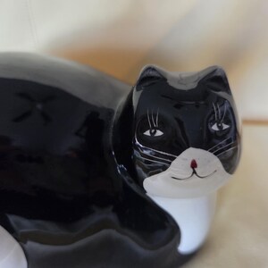 May include: A glossy ceramic cat figurine featuring a black body with a white chest and muzzle. The cat has painted black eyes with long eyelashes, a small red nose, and a subtle smile. The surface is smooth and reflective with a high-gloss finish.