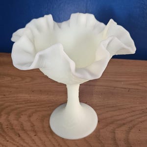 May include: A creamy off-white milk glass compote dish featuring a wide, deeply ruffled rim and a slender pedestal base. The bowl has a smooth, opaque finish with a delicate, scalloped edge that flares outward, supported by a solid, circular foot.