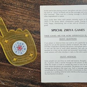 May include: A golden-yellow Ziriya game piece shaped like a hand, featuring a circular emblem with a red star design. Text on a page details the game's rules and features.