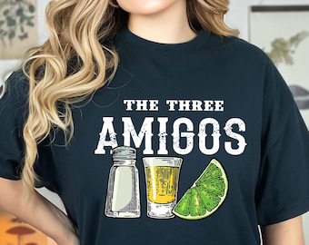 Puede incluir: Una camiseta negra con el texto "The Three Amigos" en letras blancas. La camiseta presenta un gráfico de dos saleros y un vaso de chupito con una rodaja de lima.