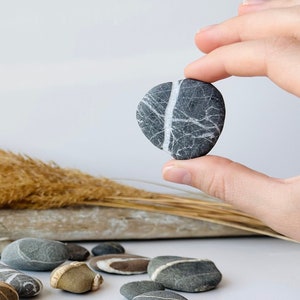 May include: A selection of smooth, grey and tan river stones with white veining. One stone is held up by a hand. Dried grasses and a piece of driftwood are in the background. The stones vary in size and shape.