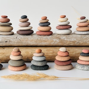 May include: A collection of twelve small stone cairns arranged in two rows on a weathered wooden log. Each stack consists of smooth, rounded river stones in various shades of grey, tan, brown, and reddish-pink, creating a balanced, natural display.