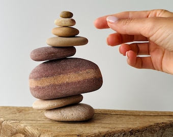 May include: A vertical stack of eight smooth, rounded river stones in varying shades of tan, beige, and reddish-brown, balanced on a weathered, textured wooden block against a plain, light-colored background.