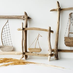 May include: Driftwood sailboat art pieces. The image shows three decorative wooden frames with small wooden sailboats inside. The sailboats have sails made of netting or wood. Dried grasses are in the foreground.