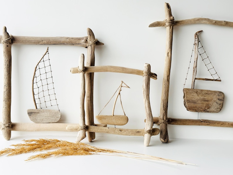May include: Driftwood sailboat art pieces. The image shows three decorative wooden frames with small wooden sailboats inside. The sailboats have sails made of netting or wood. Dried grasses are in the foreground.