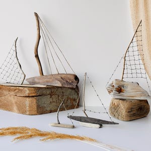 Puede incluir: Esculturas de veleros de madera flotante. Los veleros est&aacute;n hechos de madera natural y cuentan con velas hechas de red y madera. Las esculturas se exhiben sobre una superficie blanca con hierba seca.