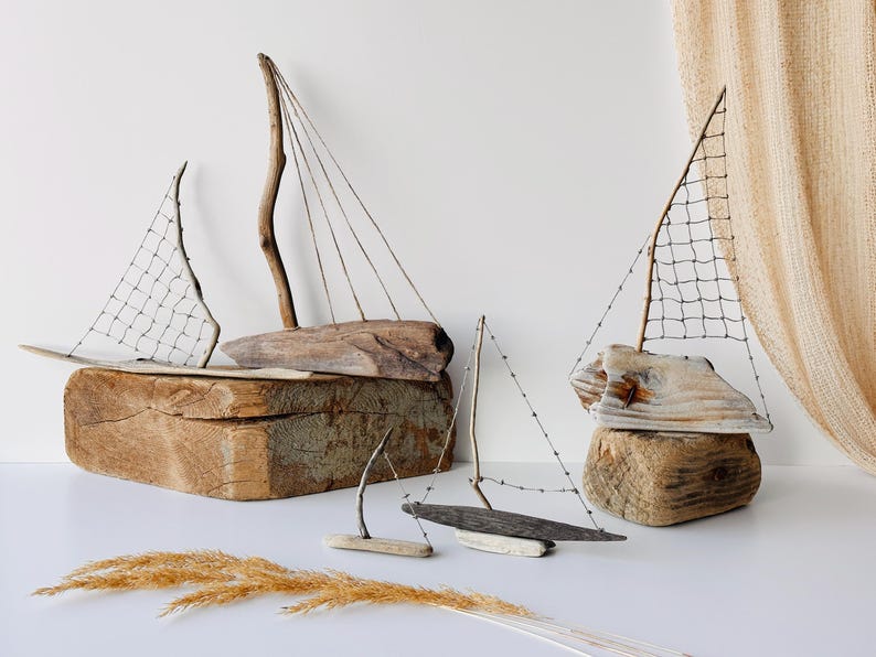 Puede incluir: Esculturas de veleros de madera flotante. Los veleros est&aacute;n hechos de madera natural y cuentan con velas hechas de red y madera. Las esculturas se exhiben sobre una superficie blanca con hierba seca.