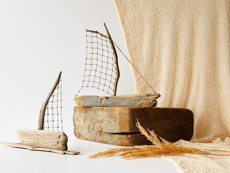 Puede incluir: Dos esculturas de veleros de madera flotante con velas de malla met&aacute;lica. Una est&aacute; sobre una base de madera, la otra sobre una base de madera flotante m&aacute;s peque&ntilde;a. El fondo incluye una tela beige y trigo seco.