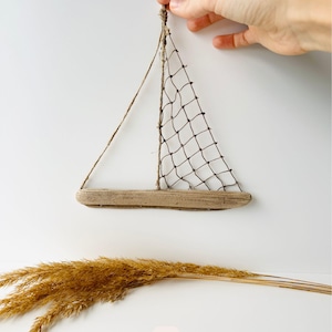 May include: A handcrafted sailboat wall hanging. The sailboat features a driftwood base, a net sail, and twine rigging. The image also includes dried ornamental grass and the number 4.