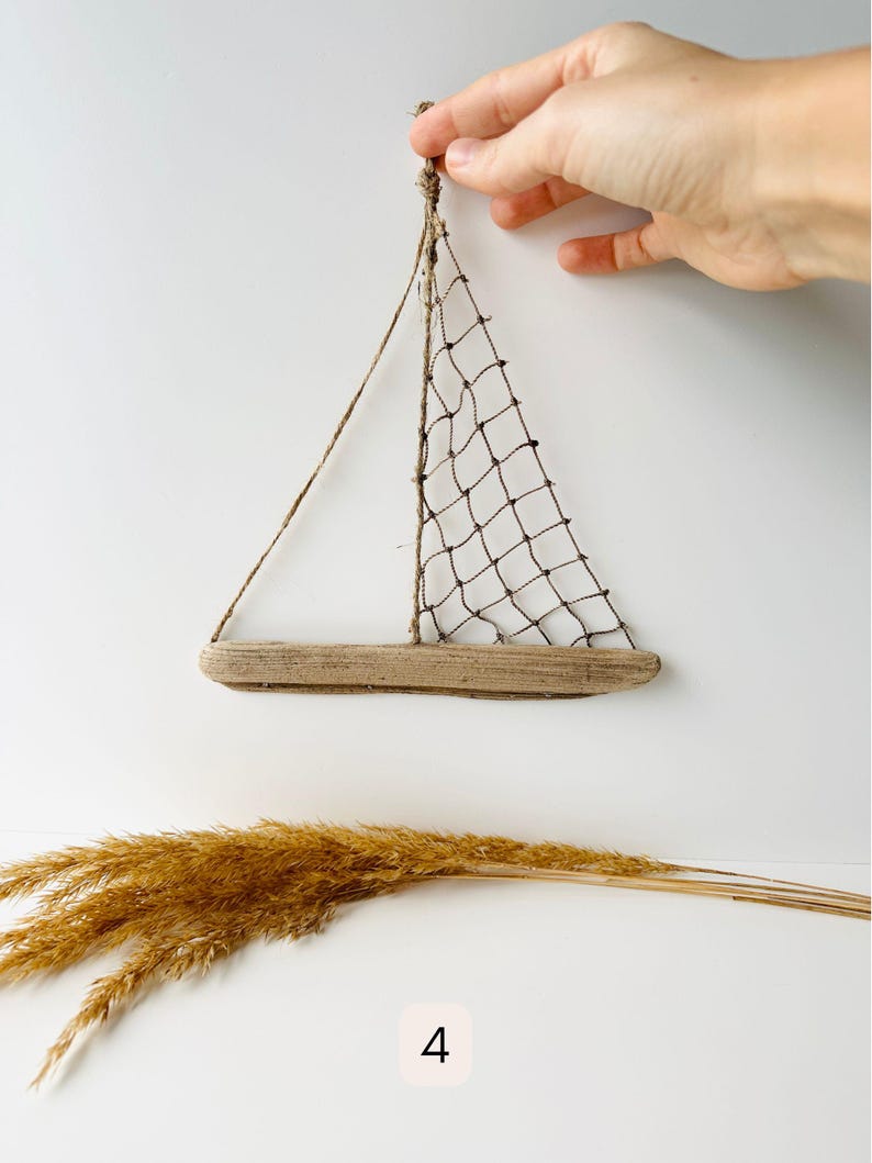 May include: A handcrafted sailboat wall hanging. The sailboat features a driftwood base, a net sail, and twine rigging. The image also includes dried ornamental grass and the number 4.