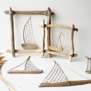 May include: Driftwood sailboat art pieces. The image shows several small sailboats made of driftwood, with white netting sails. Some are framed by driftwood rectangles. The sailboats are arranged on a white surface.