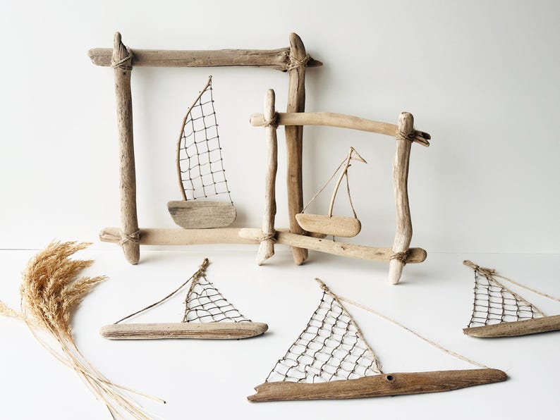 May include: Driftwood sailboat art pieces. The image shows several small sailboats made of driftwood, with white netting sails. Some are framed by driftwood rectangles. The sailboats are arranged on a white surface.