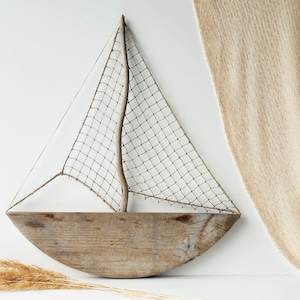 May include: A rustic wooden sailboat wall decor. The boat features a curved wooden hull, a woven net sail, and a wooden mast. The decor is a light brown color and is mounted on a white wall. Decorative dried grass is in the foreground.