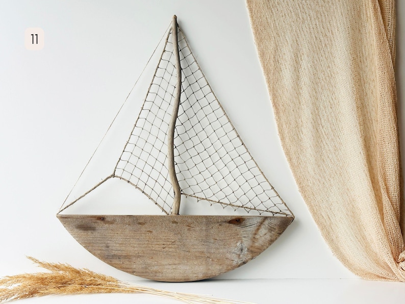 May include: A rustic wooden sailboat wall decor. The boat features a curved wooden hull, a woven net sail, and a wooden mast. The decor is a light brown color and is mounted on a white wall. Decorative dried grass is in the foreground.