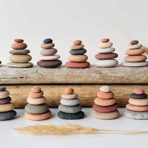 Op de afbeelding: Meerdere stapels gladde, ronde rivierstenen gerangschikt in twee horizontale rijen. De stenen variëren in kleur, waaronder tinten grijs, beige, bruin en roodachtig roze. De stapels zijn geplaatst op twee verweerde, getextureerde houten boomstammen.