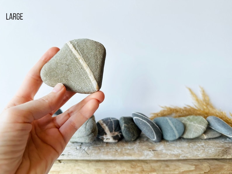 May include: A collection of smooth, gray stones with natural white and gray veining. One stone is held up, displaying a prominent white stripe. The word "LARGE" is in the upper left corner. The stones are arranged on a wooden surface.