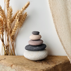 May include: A stack of five smooth, rounded stones arranged in a vertical pyramid shape. The stones vary in color, featuring shades of light gray, dark charcoal, muted mauve, pale beige, and a small dark gray stone at the very top.