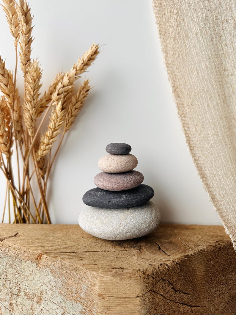 May include: A stack of five smooth, rounded stones arranged in a vertical pyramid shape. The stones vary in color, featuring shades of light gray, dark charcoal, muted mauve, pale beige, and a small dark gray stone at the very top.