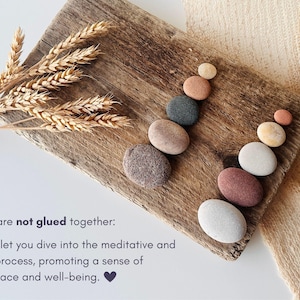 May include: Two vertical stacks of smooth, multi-colored river stones arranged on a weathered wooden board, accompanied by dried wheat stalks. The text reads: Stones are not glued together: This will let you dive into the meditative and tactile process, promoting a sense of inner peace and well-being.
