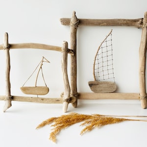 May include: Two driftwood framed wall art pieces with miniature wooden sailboats. The frames are constructed from natural, light-brown driftwood, tied together with twine. One sailboat has a wire mesh sail, the other a fabric sail. Dried wheat is in the foreground.
