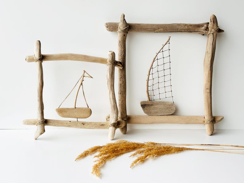 May include: Two driftwood framed wall art pieces with miniature wooden sailboats. The frames are constructed from natural, light-brown driftwood, tied together with twine. One sailboat has a wire mesh sail, the other a fabric sail. Dried wheat is in the foreground.
