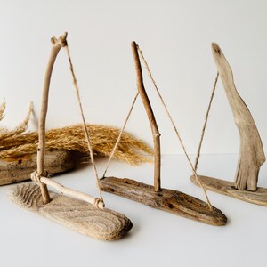 Puede incluir: Tres esculturas de madera flotante que se asemejan a veleros. Cada escultura presenta una base de madera, un soporte vertical y una pieza horizontal o en &aacute;ngulo, conectadas por hilo. La madera var&iacute;a en color de marr&oacute;n claro a oscuro. Hay hierbas secas en el fondo.