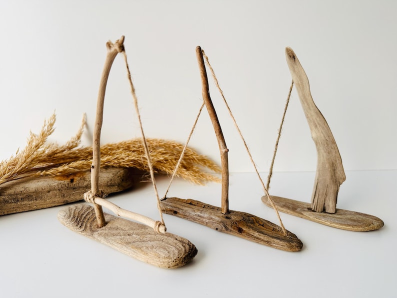 Puede incluir: Tres esculturas de madera flotante que se asemejan a veleros. Cada escultura presenta una base de madera, un soporte vertical y una pieza horizontal o en &aacute;ngulo, conectadas por hilo. La madera var&iacute;a en color de marr&oacute;n claro a oscuro. Hay hierbas secas en el fondo.