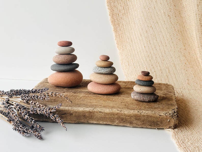 May include: Three stacks of smooth, rounded stones in varying sizes and earth tones, including shades of gray, tan, and reddish-brown. The stone towers are arranged on a rectangular piece of weathered, textured wood, accompanied by dried lavender.