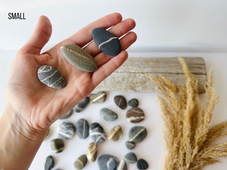 May include: A collection of small, smooth stones in various shades of gray, brown, and beige. Some stones have white or gold stripes. The word "SMALL" is in the upper left corner. The stones are displayed on a white surface.