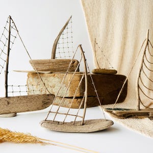 Puede incluir: Una colecci&oacute;n de veleros artesanales de madera flotante con velas de alambre y red. Los barcos var&iacute;an en tama&ntilde;o y est&aacute;n dispuestos sobre una superficie blanca con un fondo de tonos neutros, evocando un tema costero.