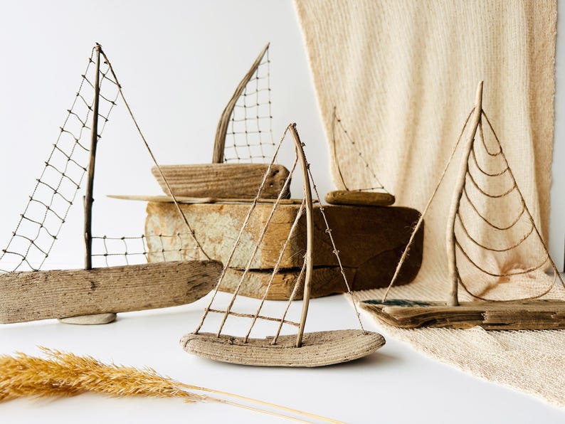 Puede incluir: Una colecci&oacute;n de veleros artesanales de madera flotante con velas de alambre y red. Los barcos var&iacute;an en tama&ntilde;o y est&aacute;n dispuestos sobre una superficie blanca con un fondo de tonos neutros, evocando un tema costero.