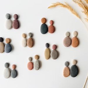 Pode incluir: Uma coleção de figuras familiares de arte em pedra em várias cores, incluindo cinza, marrom e bege, dispostas em uma superfície branca. Cada figura consiste em duas ou três pedras empilhadas para representar uma família. Caules de trigo secos no fundo.