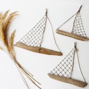 May include: Three driftwood sailboat wall hangings with netting sails and twine rigging are displayed on a white background. A bundle of dried, golden-brown ornamental grass is positioned to the left of the sailboats.