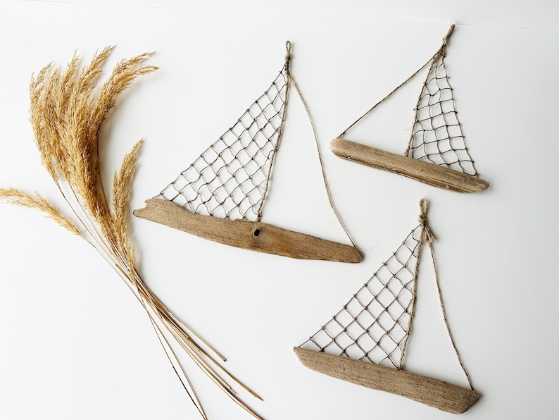 May include: Three driftwood sailboat wall hangings with netting sails and twine rigging are displayed on a white background. A bundle of dried, golden-brown ornamental grass is positioned to the left of the sailboats.